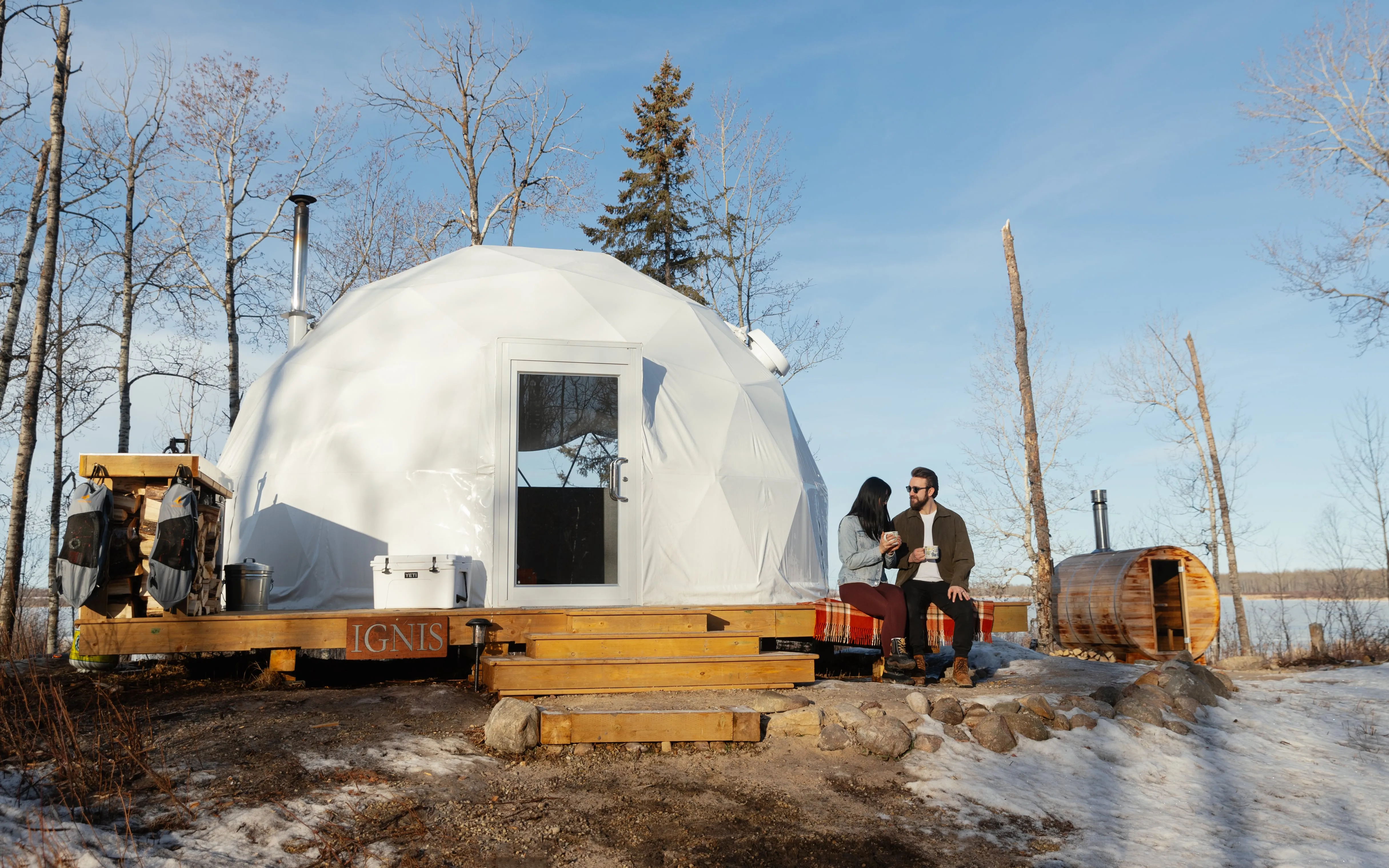 A couple sitting on a blanket on the front porch of the Ignis dome enjoying a hot cup of coffee during the winter with a view of the outdoor sauna in the background